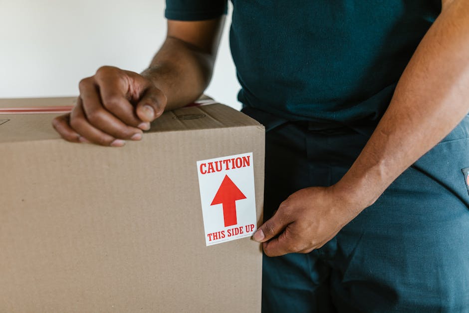 Close-up of a person in casual clothing, carefully holding a cardboard moving box with a caution label indicating 'This Side Up' and an upward arrow. The person is inside a property, preparing to load