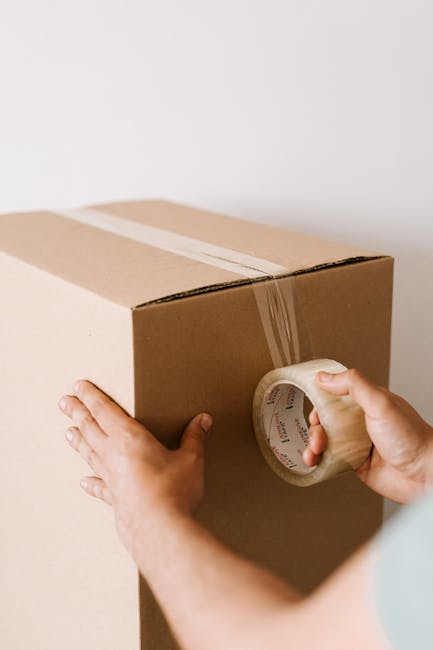 Close-up of a person in casual clothing, carefully holding a cardboard moving box with a caution label indicating 'This Side Up' and an upward arrow. The person is inside a property, preparing to load