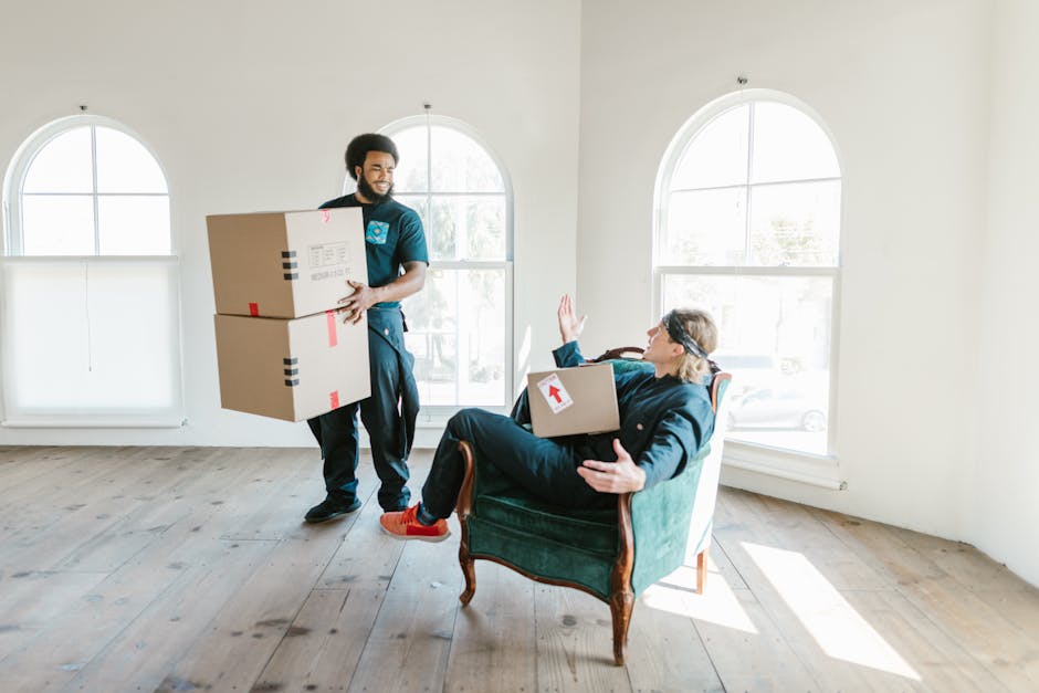 A professional removals team member is indoors, carrying two medium-sized cardboard boxes stacked one on top of the other, with red and black packing tape and labels visible on the boxes. The team mem