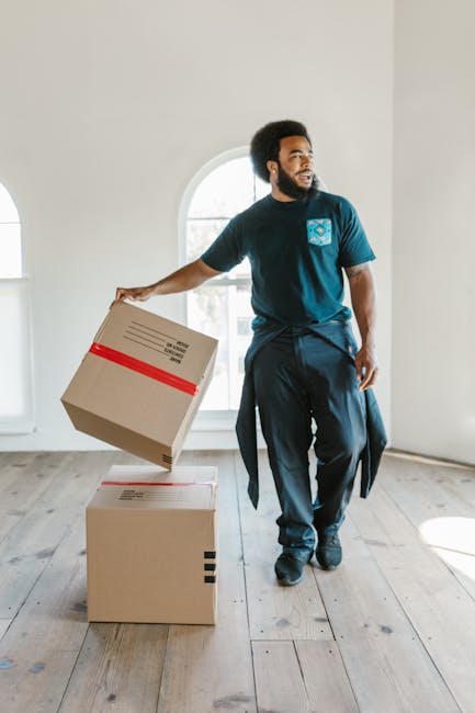 A professional removals team member is indoors, carrying two medium-sized cardboard boxes stacked one on top of the other, with red and black packing tape and labels visible on the boxes. The team mem