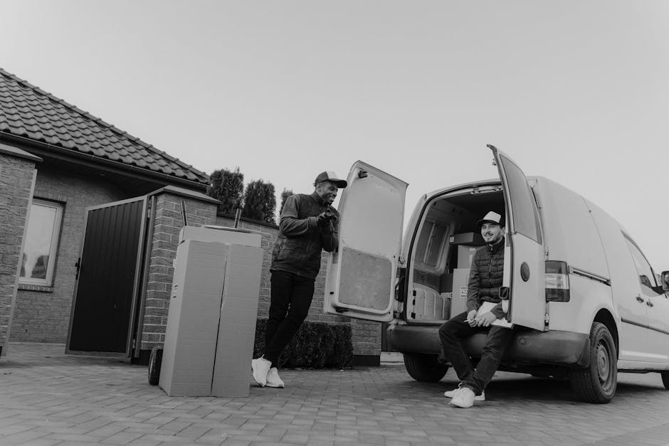 A white commercial van is parked on a grassy area next to a residential building with red brick walls and white-framed windows. The van's side panel partially shadows a tree overhead, creating a patte