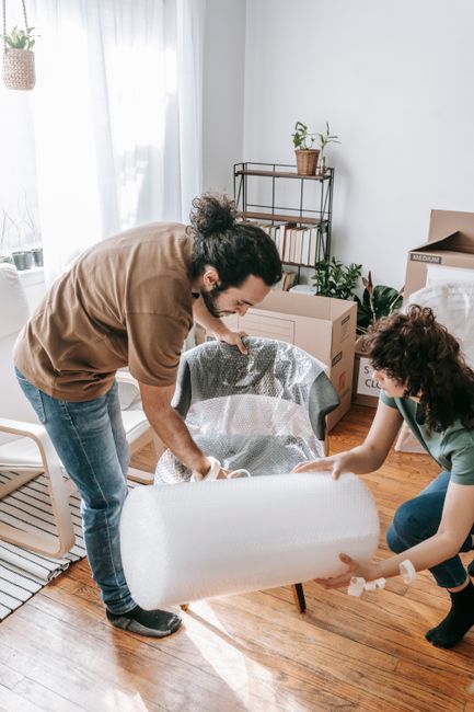 Two movers from removals company are in a well-lit living room with wooden flooring, preparing furniture for transport during a house move. The mover in the foreground, wearing a brown top and jeans,