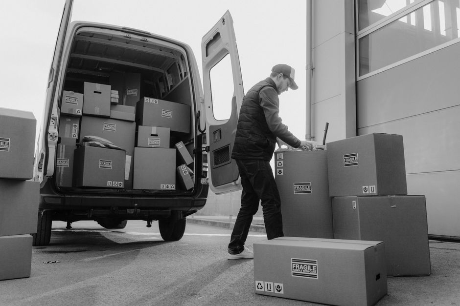 A man sitting on the open rear doors of a black van loaded with neatly stacked cardboard boxes of varying sizes, some secured with plastic wrap, in a parking lot outside a modern building with large g