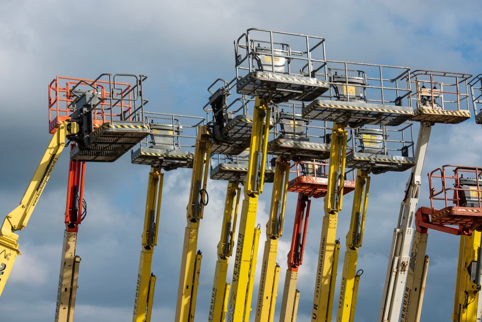 A close-up view of a yellow industrial hoist with a black hook hanging from the ceiling of a warehouse or industrial building, where overhead cranes and metal structural beams are visible in the backg