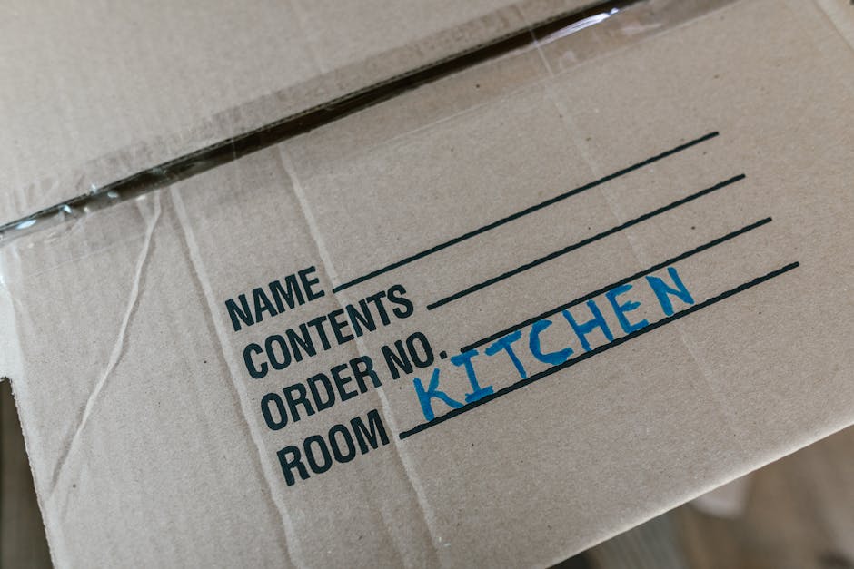 Close-up of a brown cardboard moving box with black printed labels indicating 'NAME', 'CONTENTS', 'ORDER NO.', and 'ROOM'. The 'ROOM' field is handwritten in blue ink, with the word 'KITCHEN' clearly 
